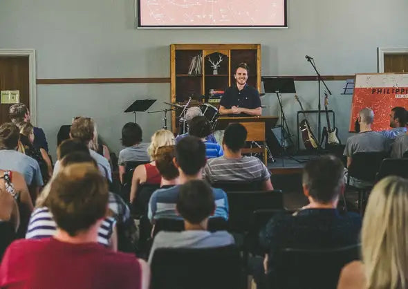 A man in a dark blue short-sleeved shirt stands behind a wooden podium, smiling toward an audience seated in rows of chairs.