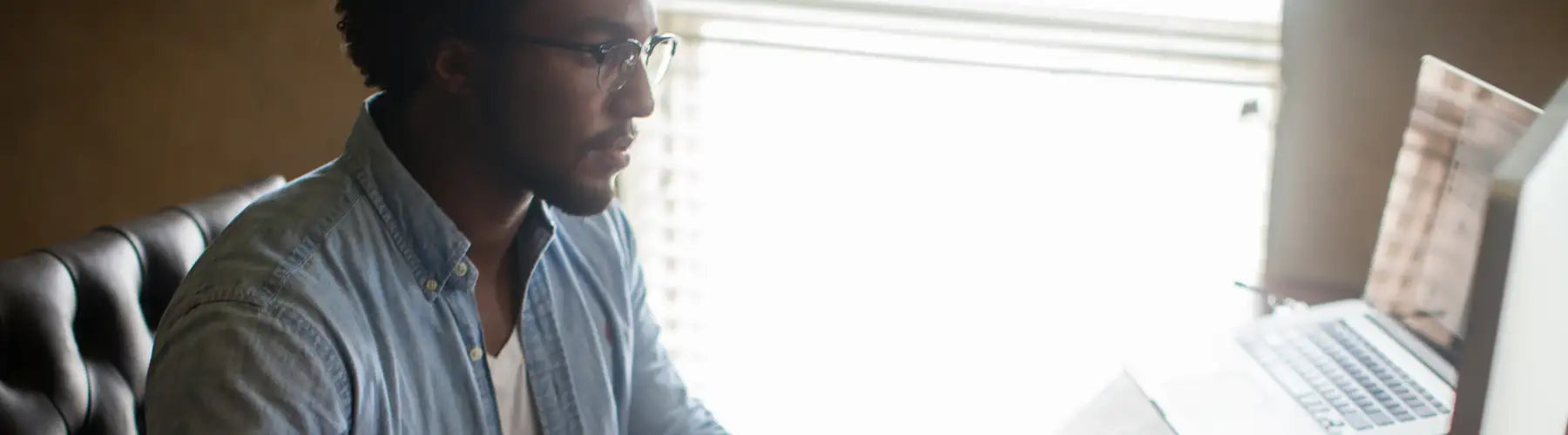 A man wearing a light blue denim shirt and glasses sits at a desk with a laptop in front of him.