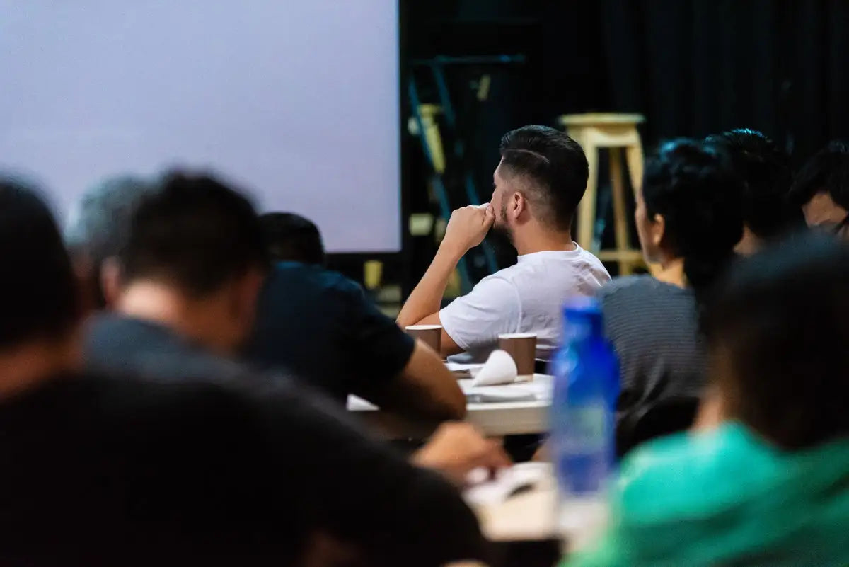 A man in a white t-shirt sits attentively at a table, his hand resting thoughtfully on his chin.