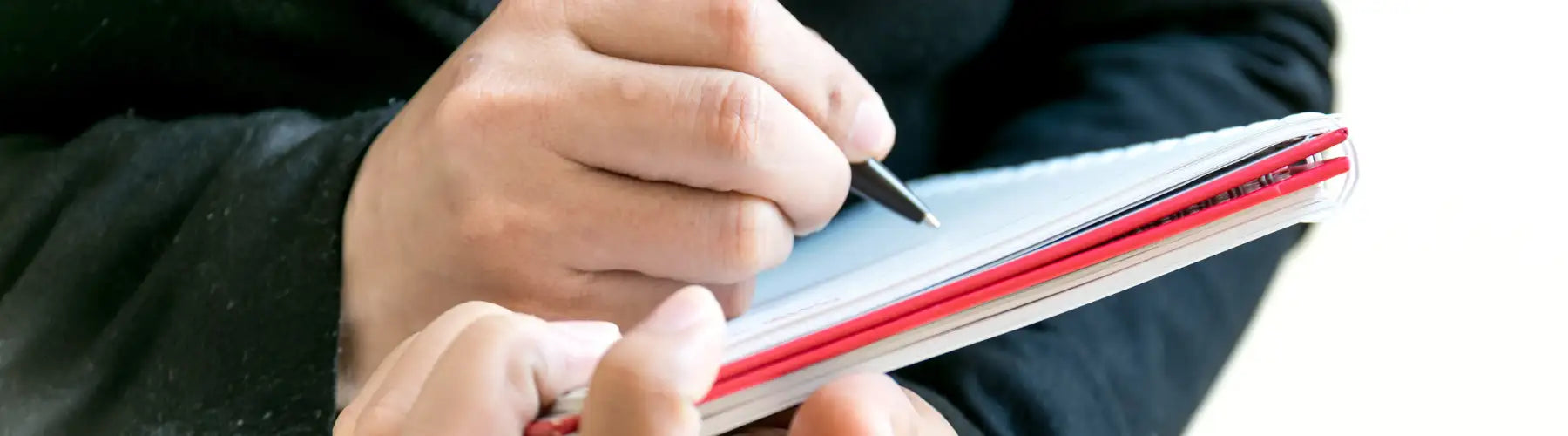 A person holds a red and white spiral-bound notebook with their hands, writing in it.