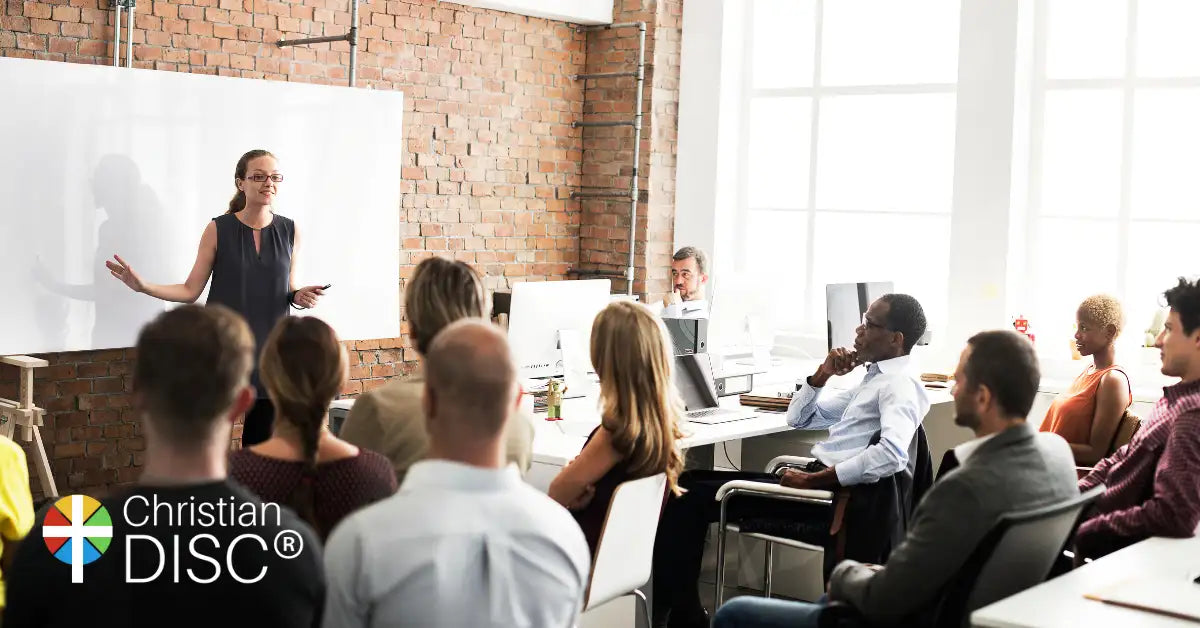 A woman in a dark sleeveless top stands before an audience, gesturing toward a whiteboard.