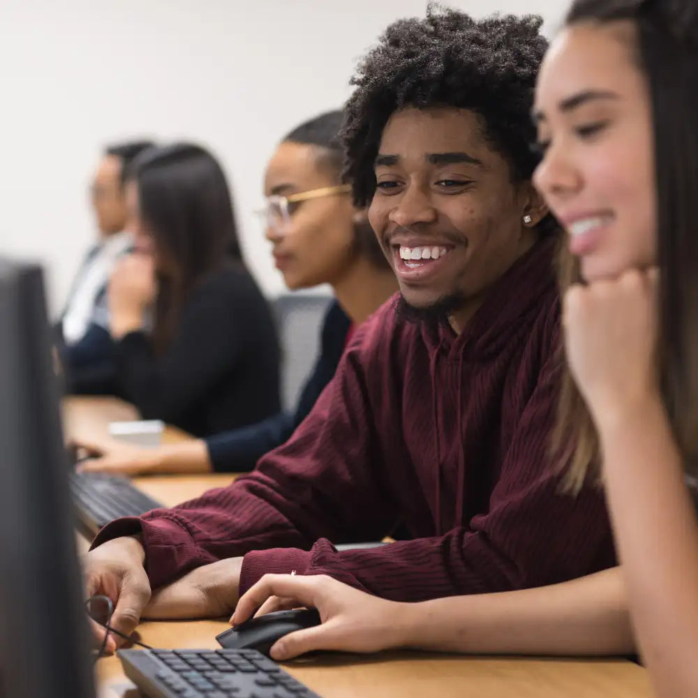 A smiling young man in a maroon ribbed hoodie sits at a desk, using a black computer mouse and keyboard.